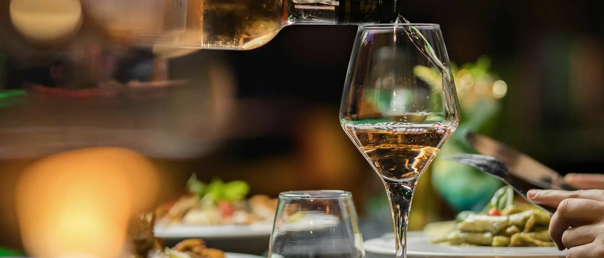 Vino blanco servido en una copa, con platillos en la mesa al fondo